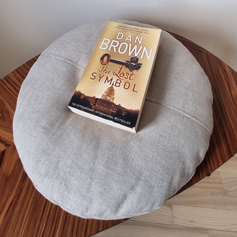Op de afbeelding: Een grijs linnen kussen met een boek getiteld "The Lost Symbol" van Dan Brown erop. De boekomslag toont een sleutel en een vrijmetselaars symbool.