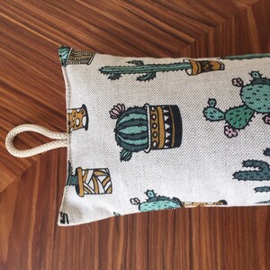 May include: A white doorstop with a cactus print. The doorstop has a rope handle and is filled with sand or beans to weigh it down.