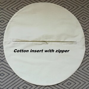 Op de afbeelding: Een ronde, witte katoenen kussenhoes met een ritsopening. De tekst "Cotton insert with zipper" is op de hoes gedrukt.