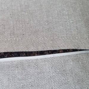 Op de afbeelding: Close-up van een grijze rits op een kussenhoes. De rits is in de stof genaaid en heeft een witte rits-trekker.
