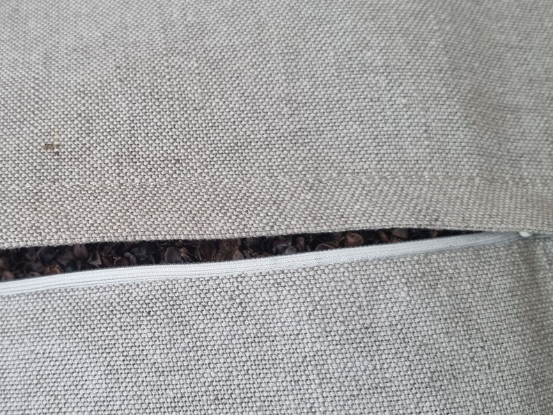 Op de afbeelding: Close-up van een grijze rits op een kussenhoes. De rits is in de stof genaaid en heeft een witte rits-trekker.