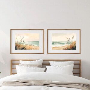 May include: Two framed watercolor paintings of a beach scene with tall grass in the foreground and a blue ocean in the background. The paintings are hung on a white wall above a bed with white bedding.