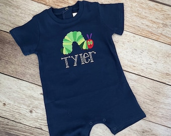 May include: A navy blue baby onesie with a green caterpillar and red apple embroidered on the front. The onesie has the name "Tyler" embroidered below the caterpillar.