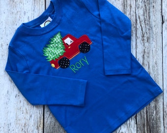 May include: A blue long-sleeved shirt with an embroidered design of a red truck carrying a green Christmas tree. The name "Rory" is embroidered below the truck.