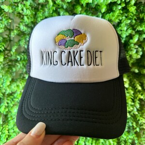 May include: A black and white trucker hat with a white front panel. The front panel features an embroidered image of a king cake and the text "KING CAKE DIET".