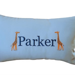 May include: A light blue pillow with the name "Parker" embroidered in navy blue. Two orange giraffes are embroidered on either side of the name.
