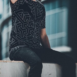 May include: Black t-shirt with a white geometric cube pattern. The shirt is paired with dark wash jeans. The person is sitting on a concrete ledge with a blurred building in the background.