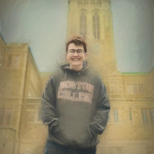 Pu&ograve; includere: Una persona con una felpa grigia del Boston College e occhiali sorride davanti a un grande edificio giallo. La felpa ha le parole "BOSTON COLLEGE" in un font rosso e bianco. Lo sfondo &egrave; un cielo nuvoloso.