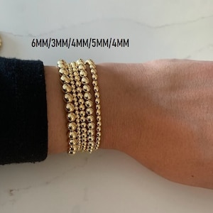May include: A set of five gold beaded bracelets stacked on a wrist. The bracelets are different sizes, with the text "6MM/3MM/4MM/5MM/4MM" visible above the bracelets.