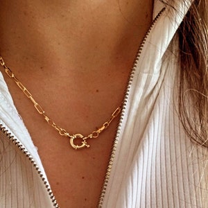 May include: A gold chain necklace with a large, ornate clasp. The necklace is worn by a person wearing a white ribbed sweater with a zipper.