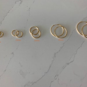 May include: A set of five gold hoop earrings in different sizes, ranging from 10mm to 35mm in diameter. The earrings are displayed on a white marble surface.