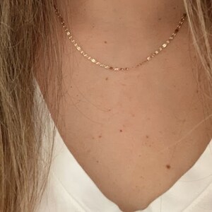 May include: A delicate gold chain necklace with a dainty, slightly textured chain. The necklace is worn by a person with a white v-neck shirt.