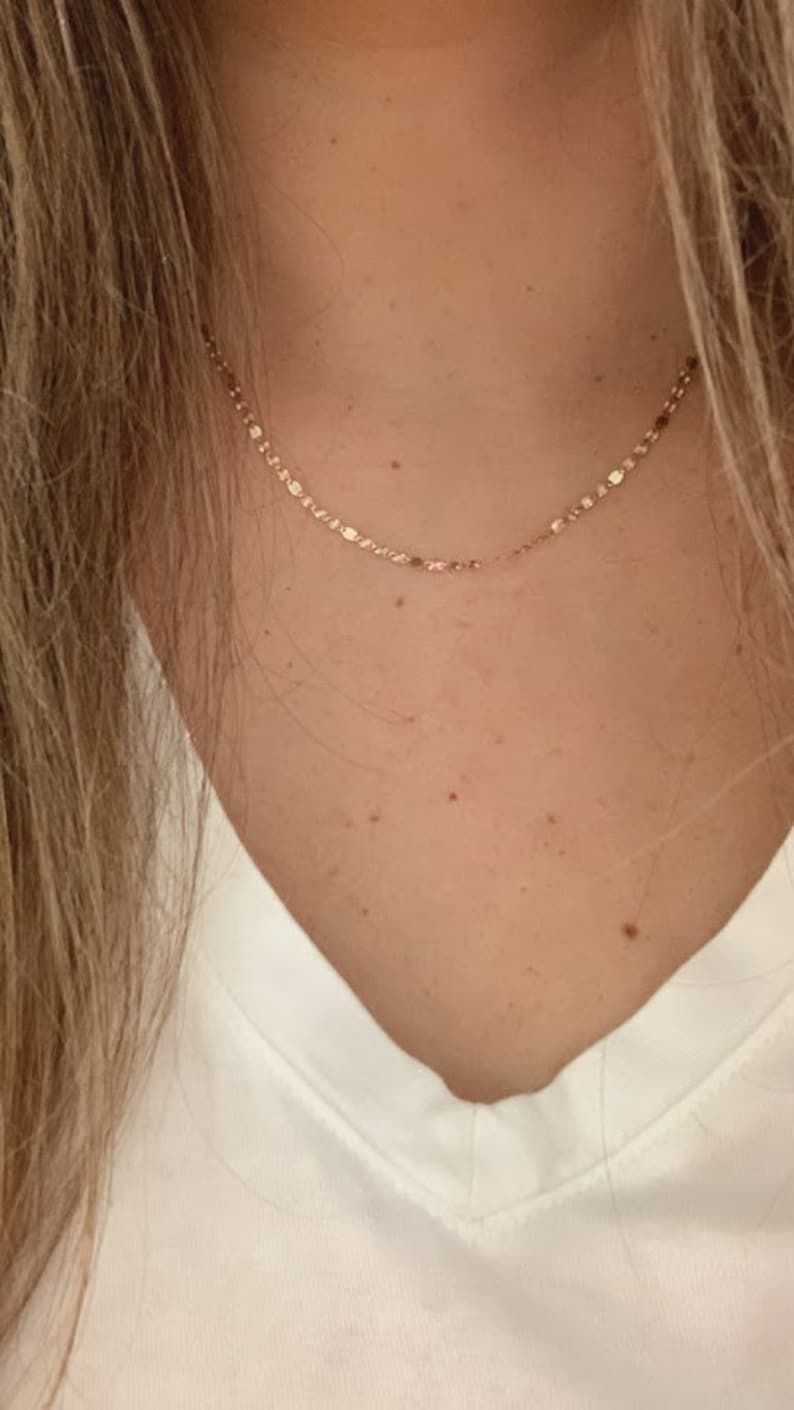 May include: A delicate gold chain necklace with a dainty, slightly textured chain. The necklace is worn by a person with a white v-neck shirt.