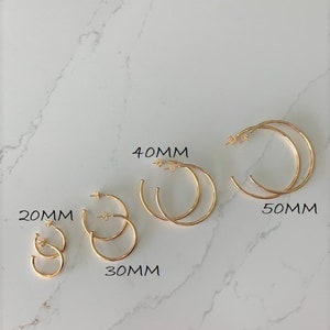 May include: Five pairs of gold hoop earrings in different sizes: 20mm, 30mm, 40mm, and 50mm. The earrings are displayed on a white marble surface.