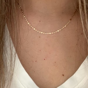 May include: A delicate gold chain necklace with a dainty, linked design. The necklace is worn by a person with a white v-neck shirt.