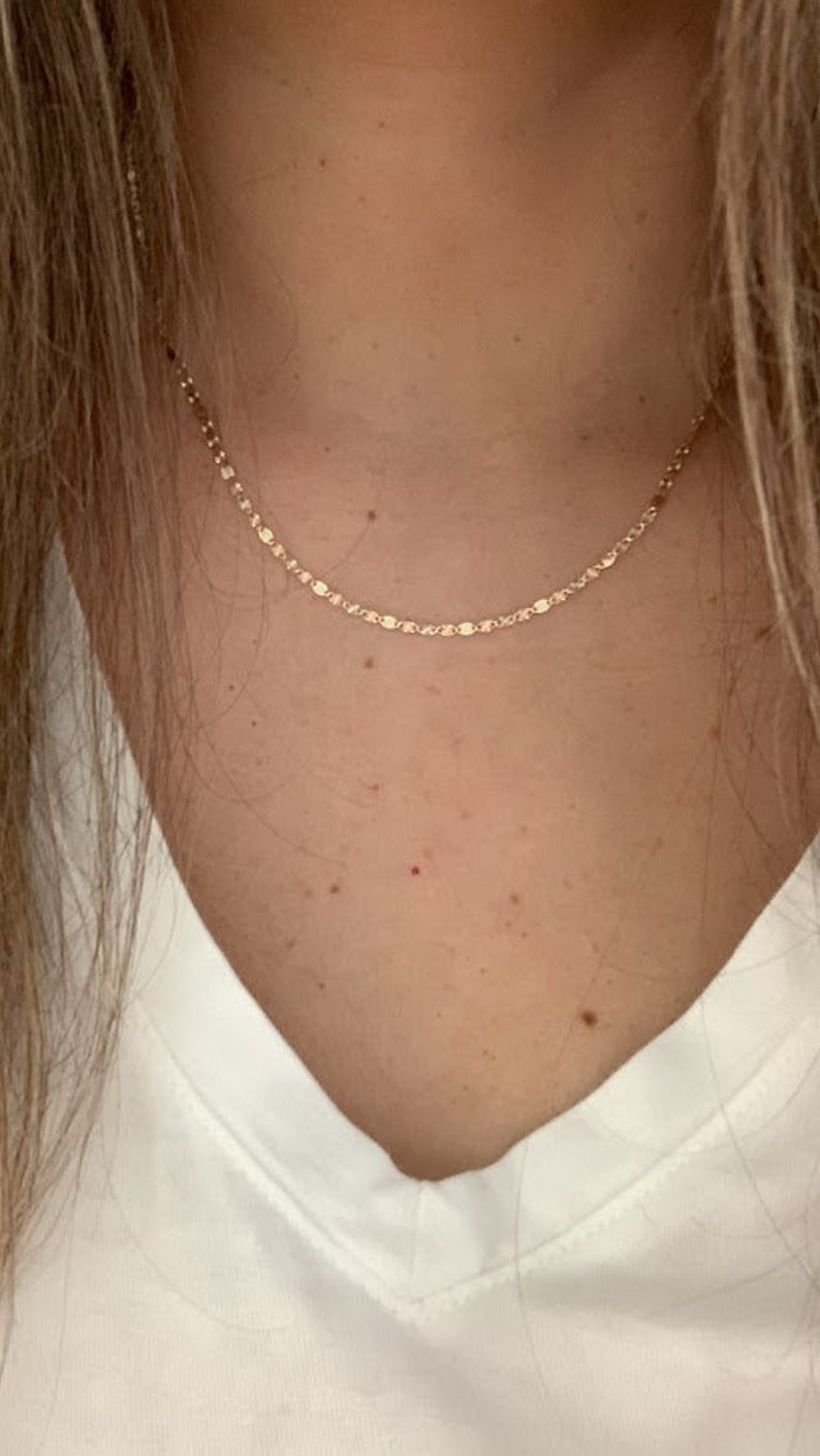 May include: A delicate gold chain necklace with a dainty, linked design. The necklace is worn by a person with a white v-neck shirt.