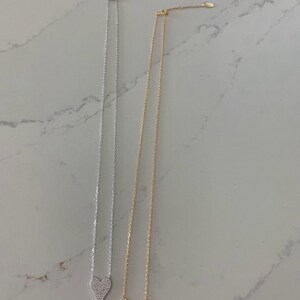 Puede incluir: Dos delicados collares con un colgante de coraz&oacute;n. El colgante de coraz&oacute;n est&aacute; cubierto de peque&ntilde;as piedras brillantes. Un collar es plateado y el otro es dorado.
