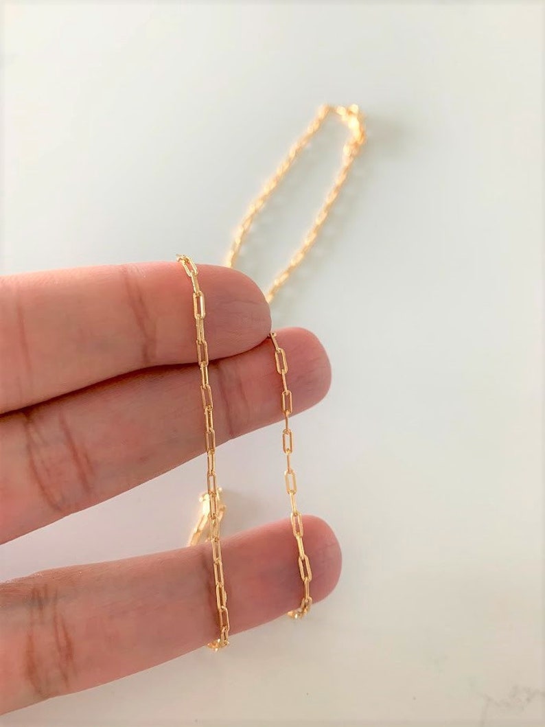 May include: A gold chain necklace with a delicate, box-style chain link. The chain is shown lying on a hand.
