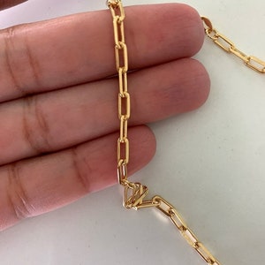 Könnte beinhalten: Eine goldene Kettenkette mit einem zarten, kastenförmigen Gliederdesign. Die Kette wird in einer Hand gezeigt, wodurch die Größe und die Details der Glieder hervorgehoben werden.
