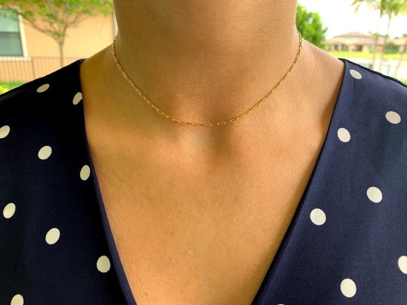May include: A delicate gold chain necklace with a simple, minimalist design. The chain is thin and features small, oval-shaped links. The necklace is worn by a person with a dark blue polka dot shirt.