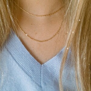 May include: A close-up of a person wearing a light blue v-neck sweater with two gold chain necklaces.