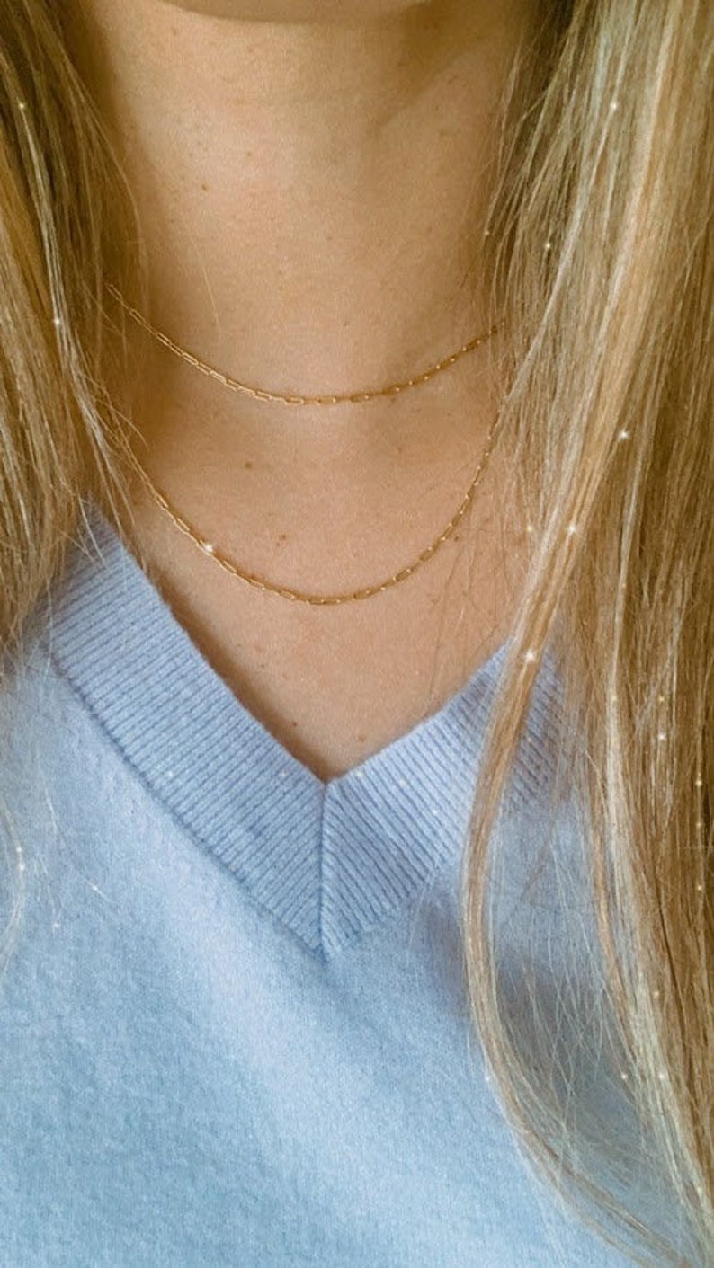 May include: A close-up of a person wearing a light blue v-neck sweater with two gold chain necklaces.