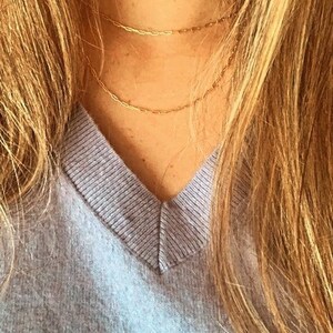 May include: A close-up of a person wearing a light purple v-neck sweater and a gold chain necklace. The necklace is layered with two chains of different thicknesses.