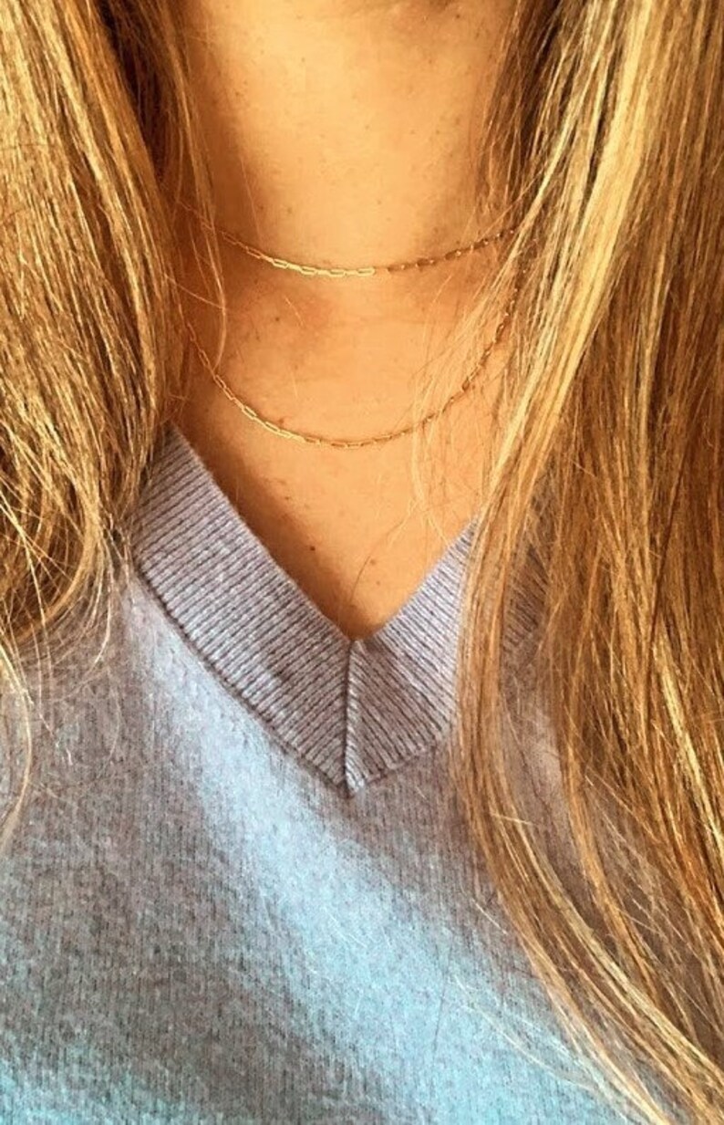 May include: A close-up of a person wearing a light purple v-neck sweater and a gold chain necklace. The necklace is layered with two chains of different thicknesses.