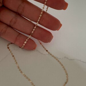 May include: A gold chain necklace with a delicate, flat oval link design. The chain is shown on a hand, with the clasp visible at the bottom.