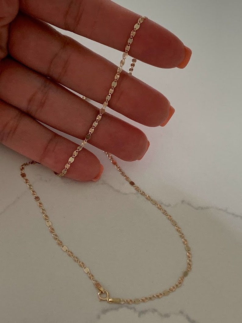 May include: A gold chain necklace with a delicate, flat oval link design. The chain is shown on a hand, with the clasp visible at the bottom.