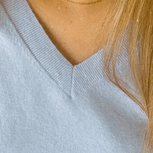 May include: A delicate gold chain necklace on a person wearing a light blue v-neck sweater.