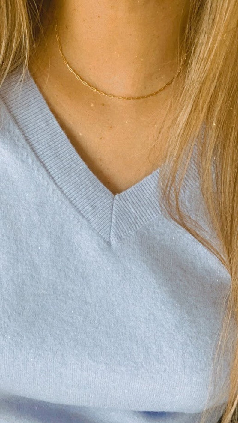 May include: A delicate gold chain necklace on a person wearing a light blue v-neck sweater.