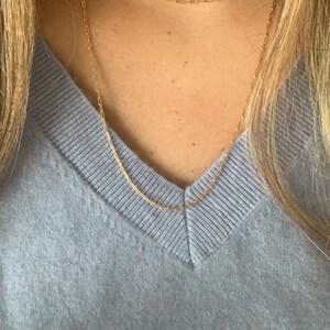 May include: A close-up of a person wearing a light blue v-neck sweater with two gold chain necklaces. The necklaces are layered, with the shorter one sitting closer to the neck.