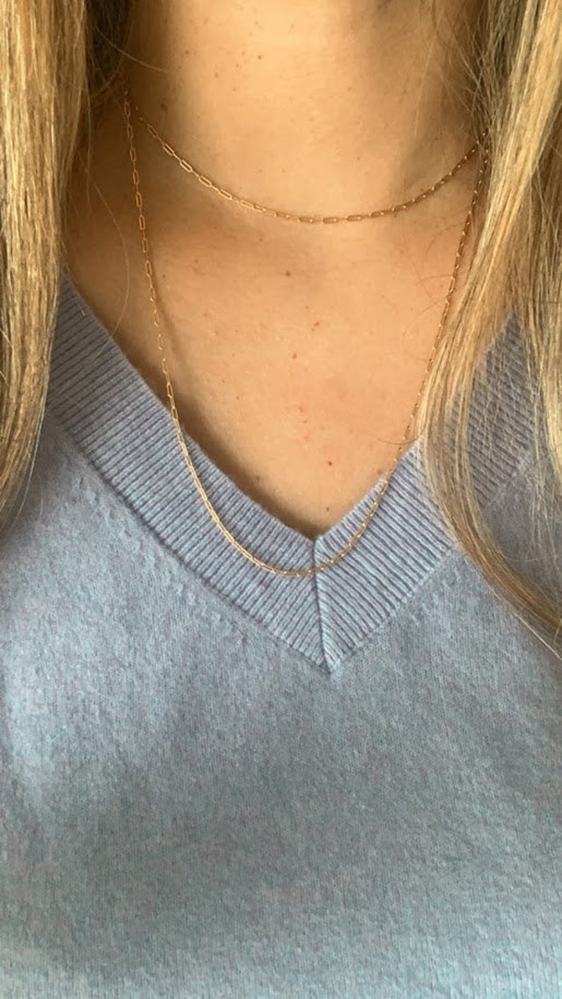 May include: A close-up of a person wearing a light blue v-neck sweater with two gold chain necklaces. The necklaces are layered, with the shorter one sitting closer to the neck.