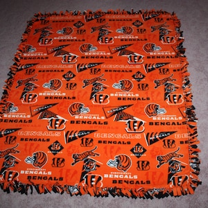 May include: An orange and black fleece blanket with the Cincinnati Bengals logo repeated throughout. The blanket has a knotted fringe border.