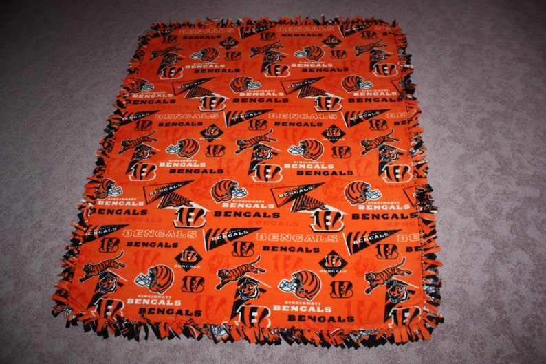 May include: An orange and black fleece blanket with the Cincinnati Bengals logo repeated throughout. The blanket has a knotted fringe border.