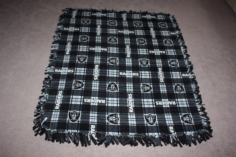 May include: Black and white fleece blanket with a plaid pattern and the word "RAIDERS" repeated throughout. The blanket has a fringe border.
