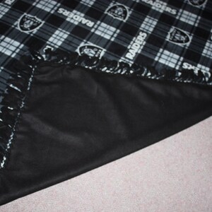 May include: Black and white fleece blanket with a plaid pattern and the word "RAIDERS" repeated throughout. The blanket has a fringe border and is folded in half.