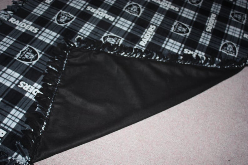 May include: Black and white fleece blanket with a plaid pattern and the word "RAIDERS" repeated throughout. The blanket has a fringe border and is folded in half.