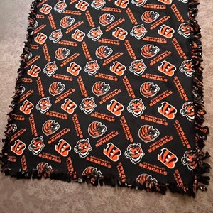 May include: A black fleece blanket with a repeating pattern of the Cincinnati Bengals logo. The logo is orange, black, and white. The blanket has a frayed edge.