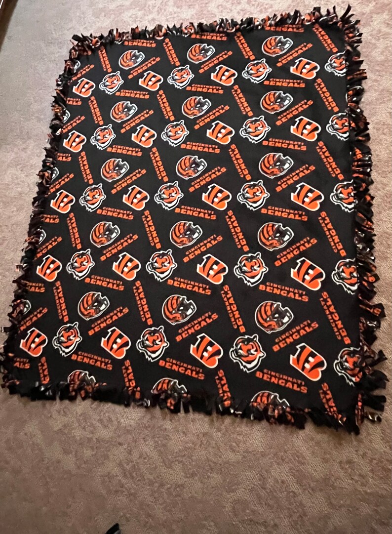 May include: A black fleece blanket with a repeating pattern of the Cincinnati Bengals logo. The logo is orange, black, and white. The blanket has a frayed edge.