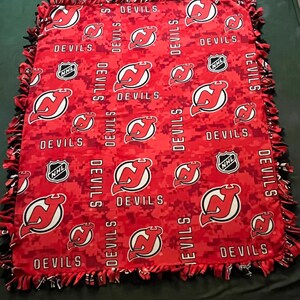 May include: Red and black fleece blanket with a camouflage pattern and the New Jersey Devils logo repeated throughout. The blanket has a knotted fringe border.