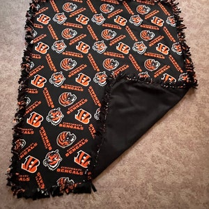 May include: Black and orange fleece blanket with a repeating pattern of the Cincinnati Bengals logo. The blanket has a knotted fringe border.