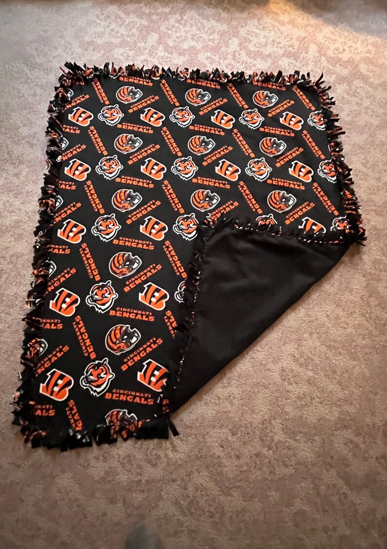 May include: Black and orange fleece blanket with a repeating pattern of the Cincinnati Bengals logo. The blanket has a knotted fringe border.