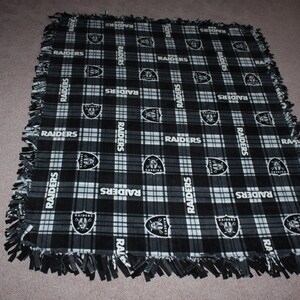 May include: Black and white fleece blanket with a plaid pattern and the word "RAIDERS" repeated throughout. The blanket has a frayed edge.