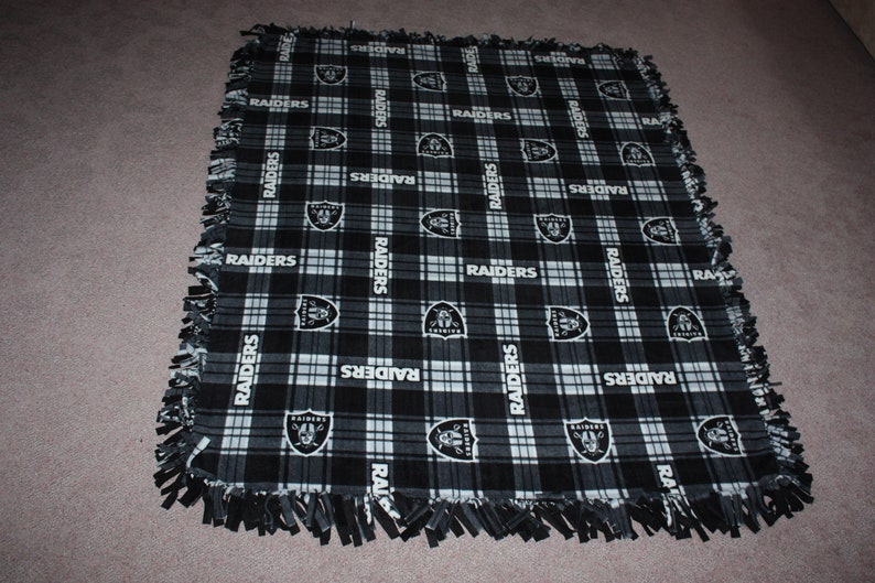 May include: Black and white fleece blanket with a plaid pattern and the word "RAIDERS" repeated throughout. The blanket has a frayed edge.