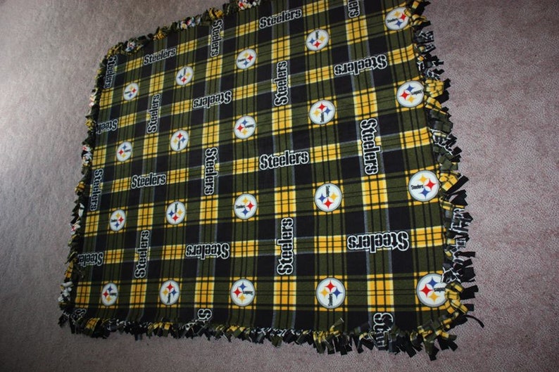May include: A black and yellow plaid fleece blanket with the Pittsburgh Steelers logo repeated throughout. The blanket has a knotted fringe border.