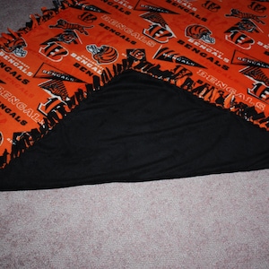 May include: A black and orange fleece blanket with the Cincinnati Bengals logo and the words "Bengals" repeated throughout. The blanket is folded in half, showing the black fleece on the inside.