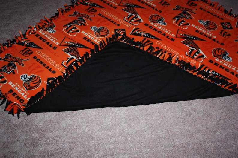 May include: A black and orange fleece blanket with the Cincinnati Bengals logo and the words "Bengals" repeated throughout. The blanket is folded in half, showing the black fleece on the inside.