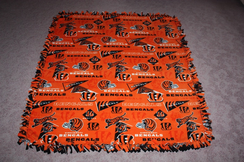 May include: An orange and black fleece blanket with a repeating pattern of the Cincinnati Bengals logo. The logo features a tiger head, the team name, and the number 16.